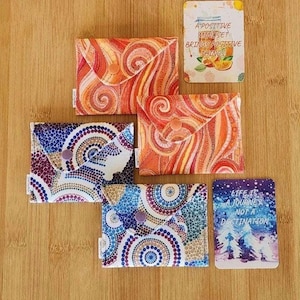 May include: An assortment of fabric wallets and small cards with inspirational quotes. The wallets feature colorful patterns in orange, blue, and purple. One card reads "A positive mindset brings positive things."
