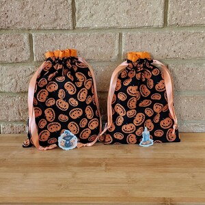 May include: Two black drawstring bags with orange pumpkin patterns. Each bag has an orange ribbon and a small sticker. The bags are on a wooden surface in front of a brick wall. Halloween themed.