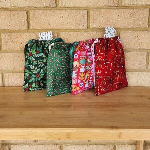 May include: Four festive drawstring gift bags with Christmas patterns. Designs include holly, candy canes, owls, and seasonal text. Each bag has a white ribbon drawstring and a tag that says "Holly Jolly".