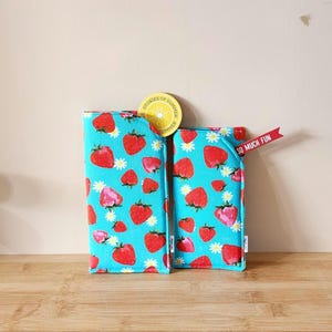 May include: Two turquoise blue fabric phone cases with a strawberry and daisy pattern. The larger case has a yellow circle sticker with the text "Stories of Summer" and a lemon slice graphic. The smaller case has a red ribbon with the text "So Much Fun".