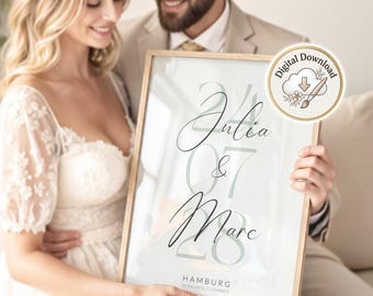 May include: A framed vertical print featuring the names Julia and Marc in elegant black script, centered over large, pale green numbers representing the date 24 07 28. The text HAMBURG and coordinates appear at the bottom with a digital download icon.