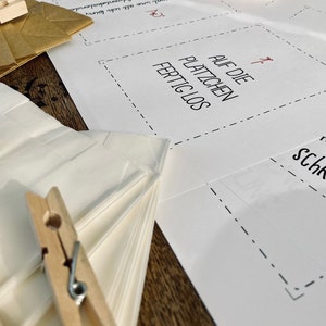 K&ouml;nnte beinhalten: Ein wei&szlig;es Blatt Papier mit dem Text "AUF DIE PLATZCHEN FERTIG LOS" darauf gedruckt. Das Papier ist in der Mitte gefaltet und es liegen zwei Holz-W&auml;scheklammern darauf.