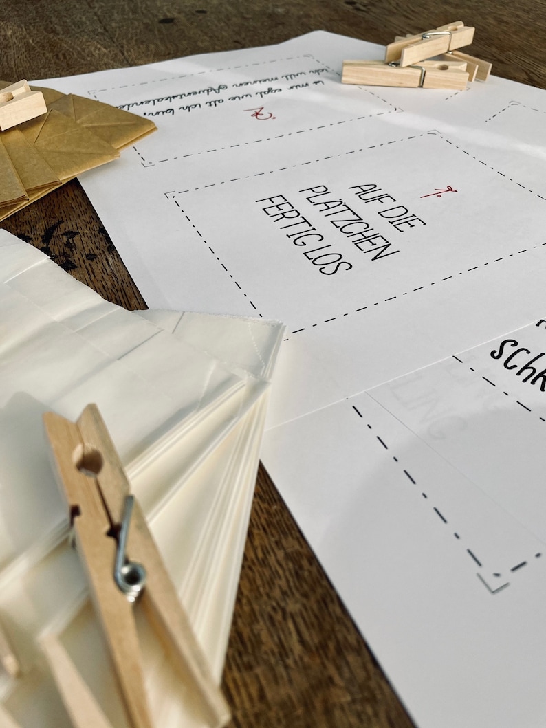 K&ouml;nnte beinhalten: Ein wei&szlig;es Blatt Papier mit dem Text "AUF DIE PLATZCHEN FERTIG LOS" darauf gedruckt. Das Papier ist in der Mitte gefaltet und es liegen zwei Holz-W&auml;scheklammern darauf.