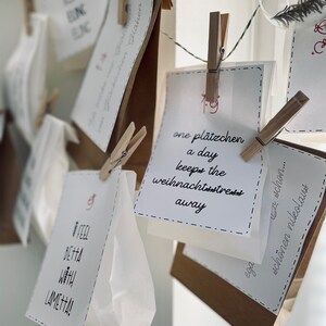 K&ouml;nnte beinhalten: Eine wei&szlig;e Schnur mit braunen Papiert&uuml;ten und wei&szlig;en Karten, die daran h&auml;ngen. Die Karten haben handschriftlichen Text, darunter "one pl&auml;tzchen a day keeps the weihnachtsstress away".