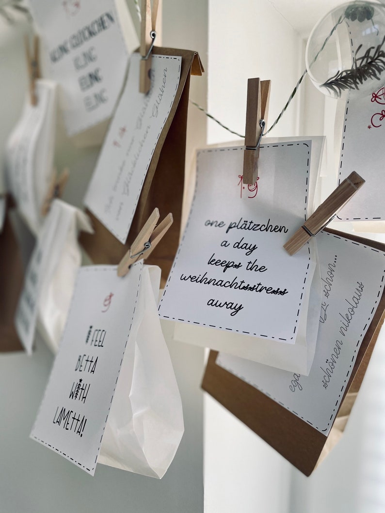 K&ouml;nnte beinhalten: Eine wei&szlig;e Schnur mit braunen Papiert&uuml;ten und wei&szlig;en Karten, die daran h&auml;ngen. Die Karten haben handschriftlichen Text, darunter "one pl&auml;tzchen a day keeps the weihnachtsstress away".