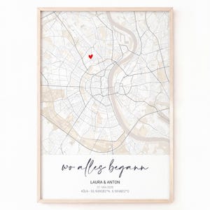 Puede incluir: Una impresión enmarcada de un mapa de Colonia, Alemania, con un corazón rojo que marca una ubicación. La impresión incluye el texto "wo alles begann" y los nombres "LAURA & ANTON", junto con una fecha y coordenadas. El marco es claro.