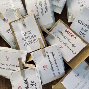 K&ouml;nnte beinhalten: Eine Sammlung kleiner brauner Papiert&uuml;ten mit wei&szlig;en Papieretiketten. Die Etiketten tragen verschiedene Weihnachtsbotschaften, die in Schwarz und Rot geschrieben sind. Einige der Botschaften lauten "Is gl&uuml;hwein o'clock", "Auf die Pl&auml;tzchen fertig los" und "Ich trinke nur Gl&uuml;hwein von Gl&uuml;cklichen".