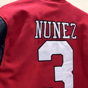 May include: Red varsity jacket with black leather sleeves. The back of the jacket has white lettering that says "NUNEZ" and the number "3" in white with a black outline.