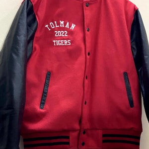 May include: A red and black varsity jacket with black leather sleeves. The jacket has a white embroidered logo that says "TOLMAN 2022 TIGERS".