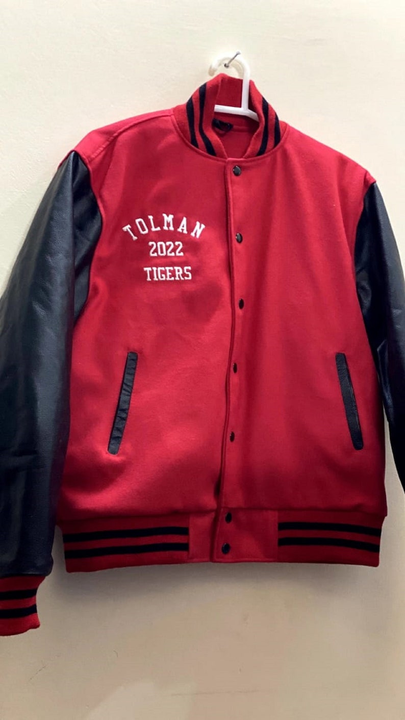 May include: A red and black varsity jacket with black leather sleeves. The jacket has a white embroidered logo that says "TOLMAN 2022 TIGERS".