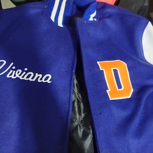 May include: A blue and white varsity jacket with the letter "D" in orange on the right side and the name "Viviana" embroidered in white on the left side.