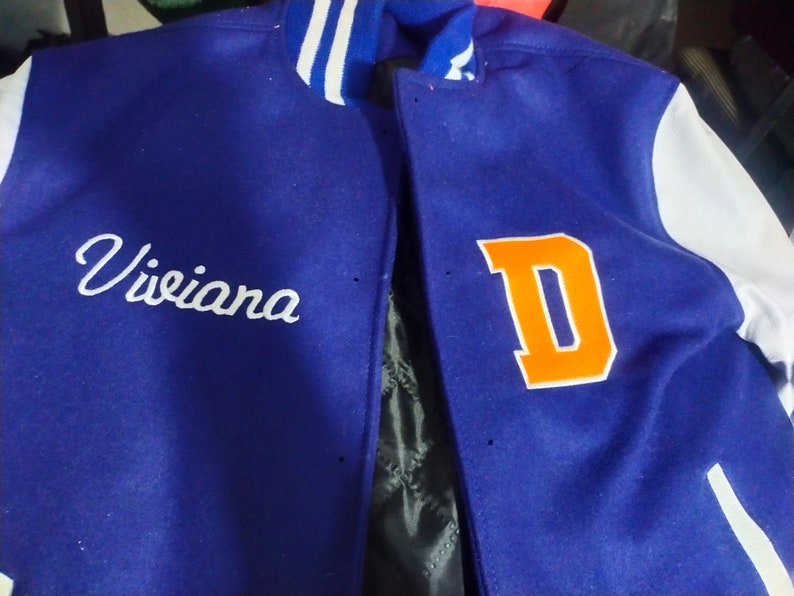 May include: A blue and white varsity jacket with the letter "D" in orange on the right side and the name "Viviana" embroidered in white on the left side.