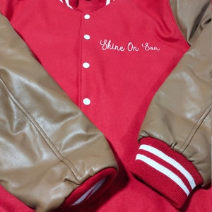 May include: Red and brown varsity jacket with white stripes on the sleeves and collar. The jacket has a button closure and the words "Shine On Son" embroidered on the front.