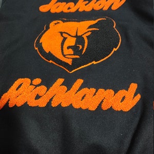 May include: Black varsity jacket with orange embroidered lettering that reads "Jackson Richland" and a black and orange bear mascot.