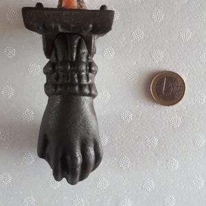 May include: A black cast iron door knocker shaped like a hand. The knocker has a detailed design and is mounted on a rectangular base.