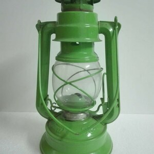 以下が含まれることがあります： 透明なガラスのグローブとワイヤーケージが付いた緑色の金属製のケロシンランタン。ランタンは両側にハンドルがあり、丸いベースがあります。