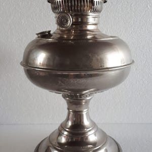 May include: A silver-coloured oil lamp with a rounded reservoir and a decorative top. The lamp features a detailed base and a chimney-like structure. A classic piece of home decor, it is a vintage item.