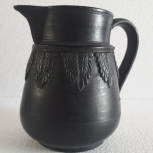 Pode incluir: Um jarro de cerâmica preta com bico e alça. O jarro tem um corpo bulboso e é decorado com um padrão de folhas repetido ao redor da parte superior. O jarro é provavelmente usado para servir bebidas.