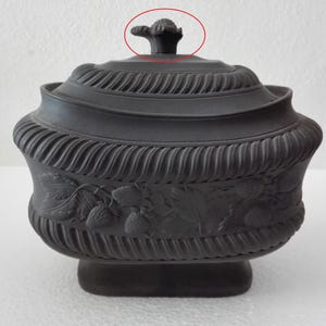 Pode incluir: Uma tigela preta com tampa, de estilo antigo, com um design floral decorativo. A tigela apresenta uma superfície detalhada e texturizada e uma tampa com uma pequena pega ornamentada. O corpo da tigela tem uma borda em relevo em forma de corda e uma base.