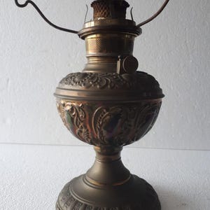 Puede incluir: Una lámpara de aceite antigua con acabado bronce. La lámpara presenta un diseño ornamentado y en relieve en el depósito y la base. Tiene una mecha y un asa metálica. La lámpara está diseñada para colocarse sobre una mesa u otra superficie plana.