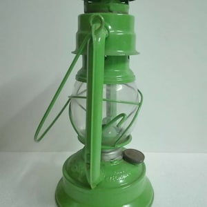 以下が含まれることがあります： 透明なガラスのグローブとワイヤーケージが付いた緑色の金属製のケロシンランタン。ランタンにはハンドルと金属製のキャップが付いています。