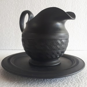 Pode incluir: Um cremoso de cerâmica preto com um padrão geométrico e um pires correspondente. O cremoso tem uma alça e um bico.