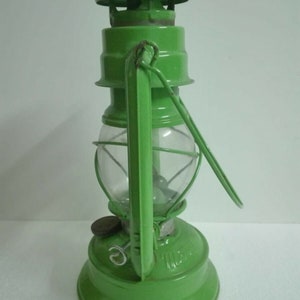 以下が含まれることがあります： ガラスのグローブとワイヤーケージが付いた緑色の金属製のケロシンランタン。ランタンにはハンドルと金属製のキャップが付いています。