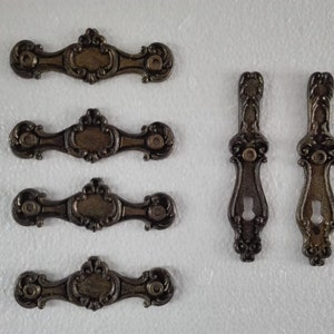 May include: Set of five antique-style metal drawer pulls. Four are ornate, rectangular pulls with a decorative center. Two are vertical pulls with a keyhole opening.