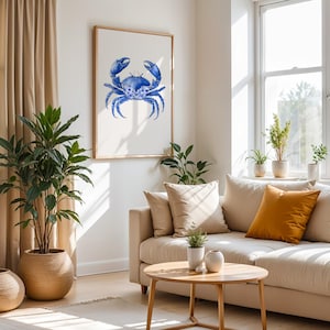 May include: A framed watercolor print of a blue crab. The artwork is displayed on a white wall above a beige sofa with decorative pillows. The room is decorated with plants and a wooden coffee table.