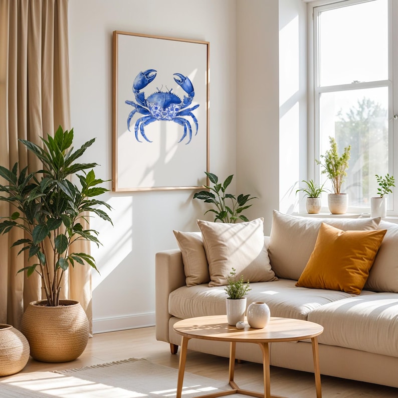 May include: A framed watercolor print of a blue crab. The artwork is displayed on a white wall above a beige sofa with decorative pillows. The room is decorated with plants and a wooden coffee table.