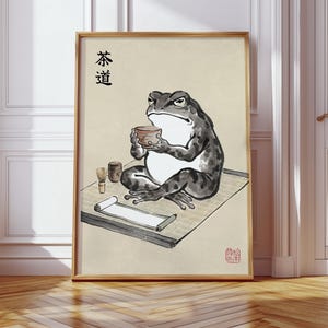 May include: Framed art print featuring a frog in a traditional Japanese tea ceremony. The frog is holding a tea bowl, with a tea whisk and scroll nearby. The artwork is in shades of black, gray, and brown, with Japanese text in the upper left corner.