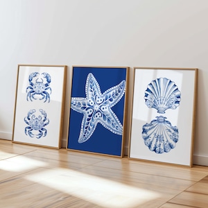 Puede incluir: Tres impresiones enmarcadas con ilustraciones azules y blancas de una estrella de mar, un cangrejo y una concha. Las impresiones están sobre un fondo blanco y tienen un marco de madera claro.