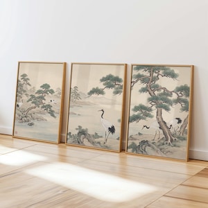 May include: Three framed art prints featuring traditional Japanese landscape scenes. Each print depicts a crane, trees, and mountains in muted tones of beige, green, and gray. The frames are a warm gold color.