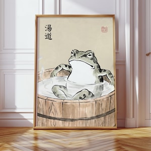 May include: A framed print featuring a frog relaxing in a wooden tub. The frog is gray and white, with a stern expression. The tub is filled with water, and the background is a light beige. Japanese text is visible in the upper left corner.