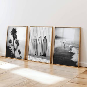May include: A triptych of framed black and white prints with a coastal theme. The images show palm trees, surfboards, and surfers in the sea. The frames are light brown. The prints are displayed on a wooden floor.