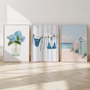 Peut inclure: Trois impressions aquarelles encadrées. L'impression de gauche présente des hortensias bleus dans un bocal en verre. L'impression du centre montre un bikini bleu suspendu à une corde à linge sur des rayures bleues et blanches. L'impression de droite représente des cabanes de plage.