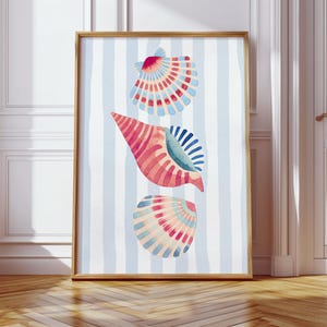 May include: A framed watercolour print depicting three seashells in red, pink, blue, and peach hues. The seashells are set against a backdrop of vertical blue and white stripes. The frame is made of light brown wood.