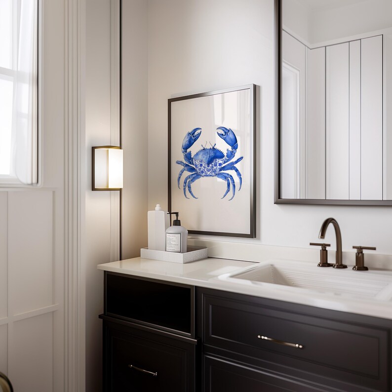 May include: A framed watercolor print of a blue crab hangs above a bathroom vanity. The crab art is displayed with a white tray holding a soap dispenser and a bottle. The vanity is dark with a white countertop.