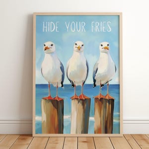 Op de afbeelding: Een ingelijste kunstprint met drie witte meeuwen met oranje snavels en poten, staand op houten palen tegen een blauwe oceaanachtergrond. De tekst HIDE YOUR FRIES staat in witte hoofdletters bovenaan de illustratie afgebeeld.