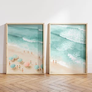 Pode incluir: Duas impressões de arte emolduradas com uma cena de praia. As impressões apresentam uma representação em aquarela de uma praia arenosa com água turquesa, ondas e pessoas. Guarda-sóis rosa e azuis fornecem sombra para os banhistas.