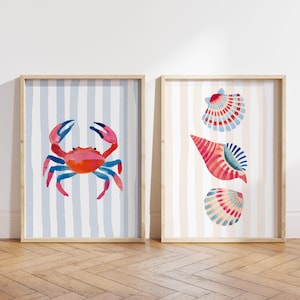 May include: Two framed watercolor prints. One features a red and blue crab on a blue and white striped background. The other print displays three seashells in red, blue, and white on a beige and white striped background. Both are in wooden frames.