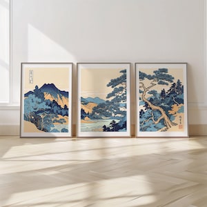 May include: Three framed art prints featuring Japanese landscape scenes. Each print showcases a different view of mountains, trees, and water, rendered in shades of blue, beige, and brown. The artwork is displayed on a light-colored wall.