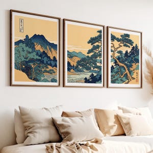 May include: Three framed landscape prints in a Japanese style. Each print depicts mountains, trees, and water in blue, green, and yellow hues. The frames are a warm brown, and the prints are displayed above a bed with neutral-toned pillows.