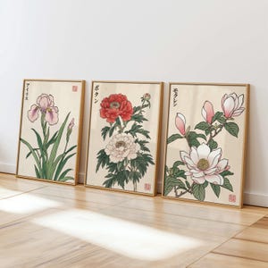 May include: Three framed botanical prints featuring Japanese-style illustrations of flowers. The prints showcase an iris, a peony, and a magnolia, each with detailed depictions of petals and leaves. The frames are a light brown color, and the background is a cream color.