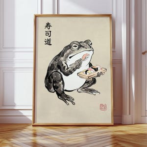 Könnte beinhalten: Gerahmter Kunstdruck mit einer Aquarellillustration eines Frosches, der Sushi isst. Der Frosch, in Grau- und Schwarztönen, hält Essstäbchen und ein Holztablett mit Sushi. Japanischer Text ist sichtbar.