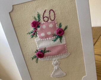 May include: A handmade card with a pink and white cake with a number 60 on top. The cake is decorated with pink flowers and green leaves. The card is on a wooden surface.