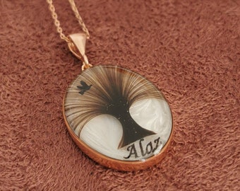May include: A silver necklace with a white oval pendant featuring a black silhouette of a tree with a bird on a branch. The name "Alay" is inscribed on the pendant.