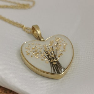 Op de afbeelding: Een gouden ketting met een hartvormige hanger. De hanger is gevuld met helder hars en bevat een gedroogd bloemenboeket met gouden accenten.