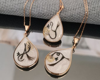 May include: Three gold necklaces with teardrop-shaped pendants. Each pendant features a delicate black and white design encased in clear resin.