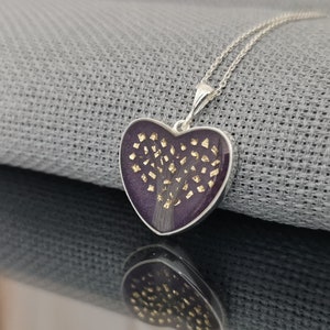Op de afbeelding: Een zilveren hartvormige ketting met een hanger van paarse hars met een gouden boomontwerp.